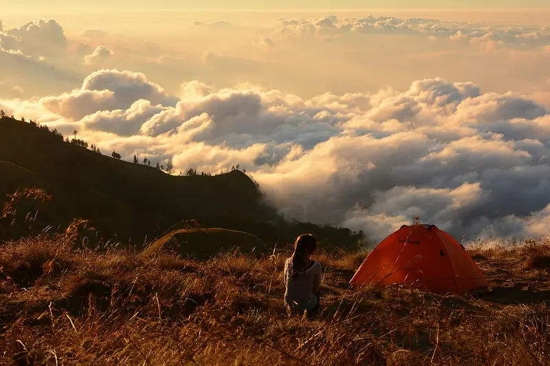 A hiker beside an orange tent above a sea of cloud on Mount Rinjani